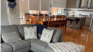 A modern living room with a grey sectional sofa, wooden dining chairs, and a kitchen area with a bar stool.