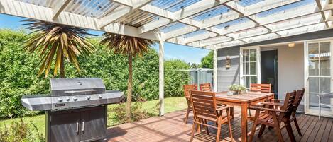 A patio with a grill, wooden table, and chairs, under a glass roof.