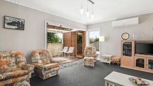 Sala de estar con dos sillones estampados, una mesita, un mueble con televisor y un ventanal corredizo que da a un área exterior con terraza de madera y sillas.