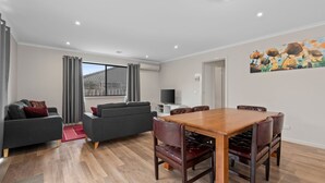 Un salon moderne avec un canapé gris, une table à manger en bois avec des chaises en cuir, un téléviseur à écran plat et une grande fenêtre avec des rideaux.