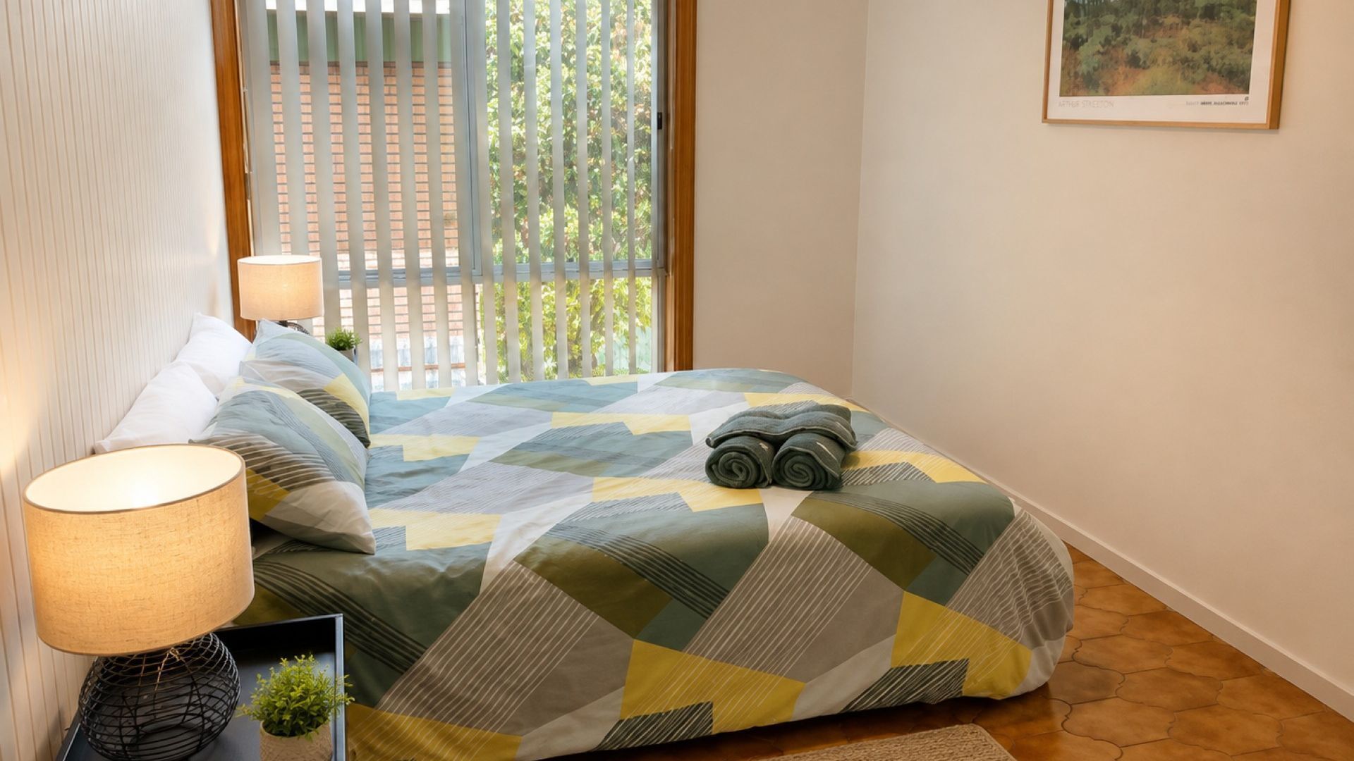 Een opgemaakt bed met een bedspreider met geometrisch patroon, een bedlamp en een lijst met een foto aan de muur.