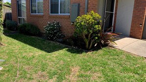 A residential property with a brick facade, a small front yard with a lawn, and a variety of shrubs and small trees.