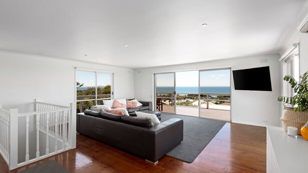 A spacious living room with a large gray sofa, a white railing, and a view of the ocean.