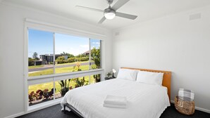 A bedroom with a large window, a bed, a ceiling fan, and a view of a house and garden.