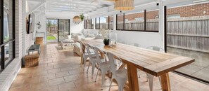 Une vaste terrasse dotĂ©e dâune table en bois et de chaises blanches, dâun luminaire suspendu et offrant une vue sur le jardin Ă travers de grandes fenĂȘtres.