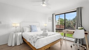 Une chambre à coucher comprenant un lit, des tables de chevet, un bureau, une chaise, un ventilateur de plafond et une grande fenêtre donnant sur l’extérieur.