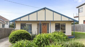 Une maison de plain-pied avec un toit à pignon, un porche à l’avant et un petit jardin devant, agrémenté d’arbustes et d’une pelouse.