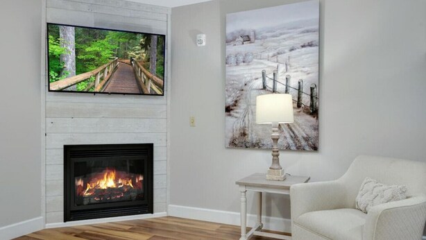 Ein Wohnzimmer mit Kamin, einem darüber hängenden Fernseher, einem Gemälde an der Wand, einer Lampe auf einem Beistelltisch und einem weißen Sessel.
