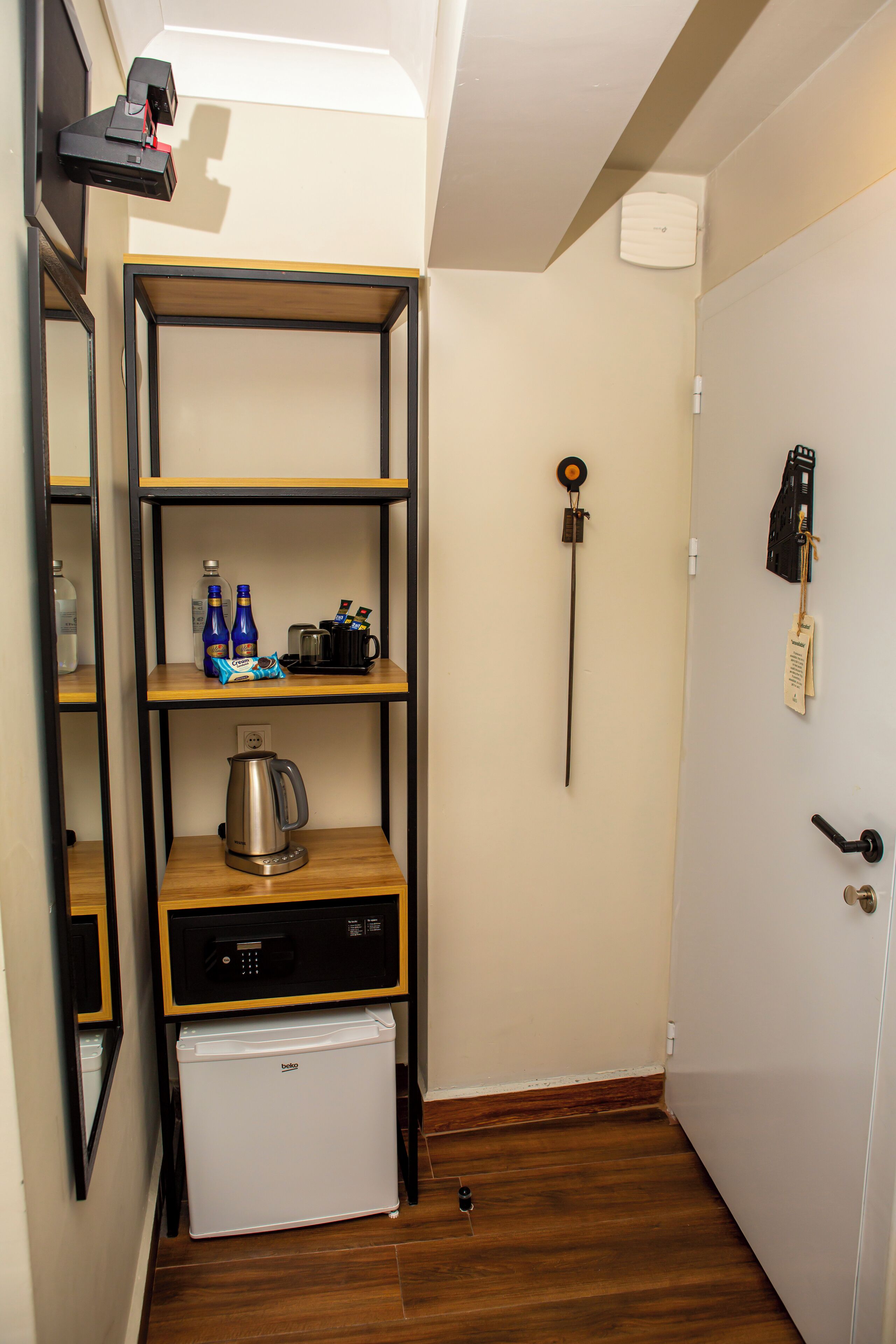 Una cocina compacta con un refrigerador, una cafetera y un hervidor sobre la encimera. Hay repisas arriba que sostienen botellas y una máquina de café.
