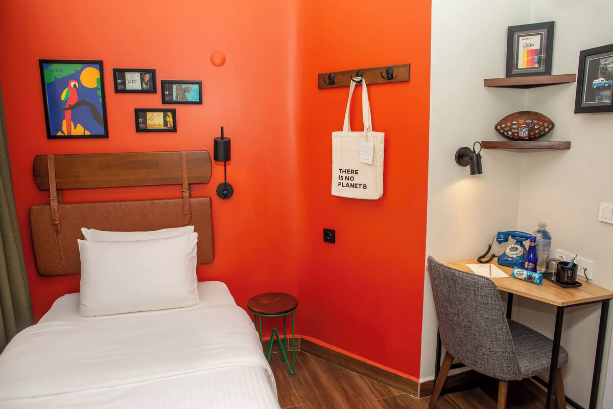 A hotel room with a bed, a desk, and a chair. There are framed pictures on the wall and a tote bag with text on it.