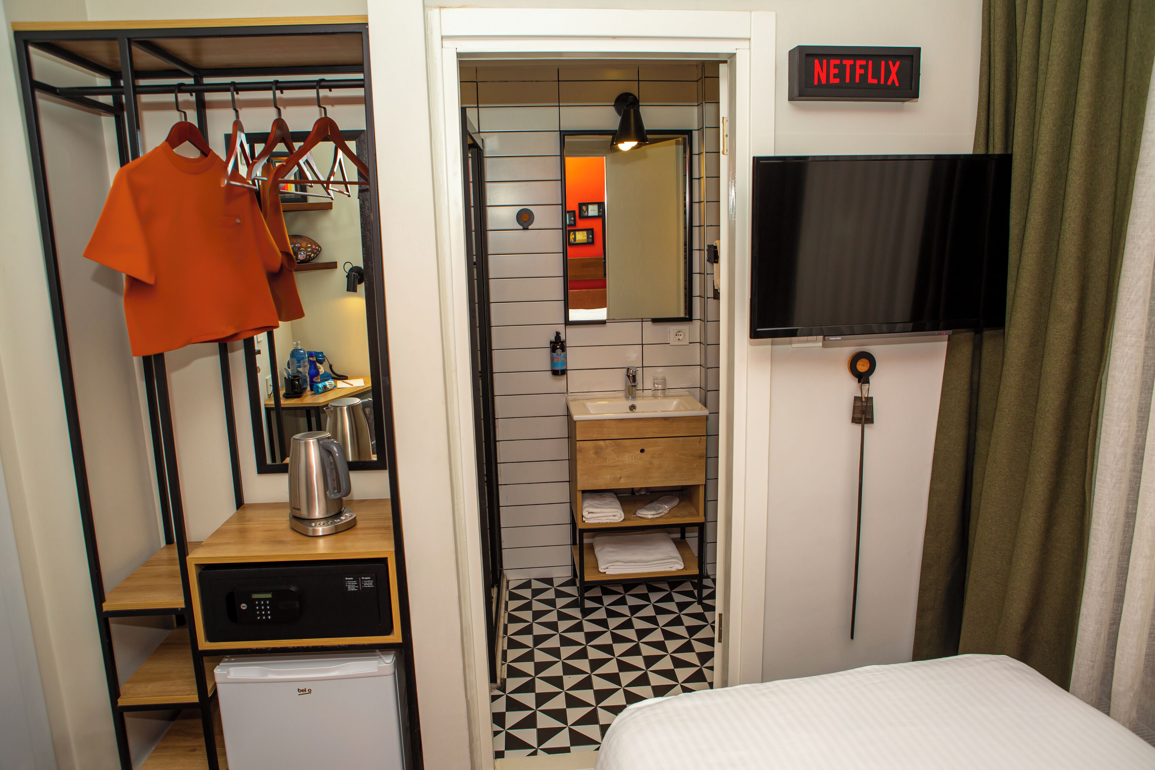 A compact hotel room with a bed, a small kitchenette, a bathroom with a sink and mirror, and a TV mounted on the wall.