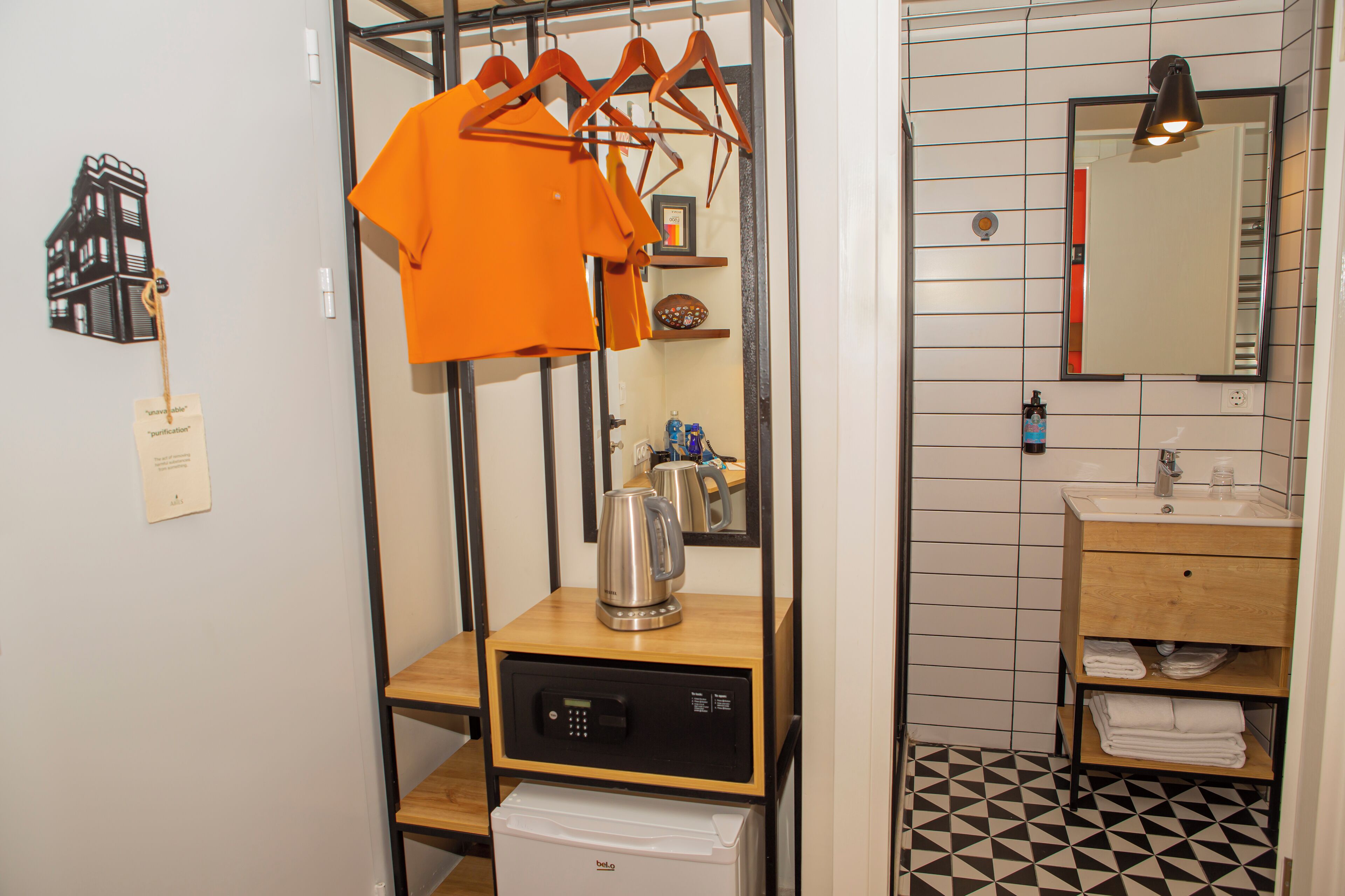 A compact hotel room with a kitchenette, a wooden shelf, a hanging rack with clothes, a mirror, and a tiled floor.
