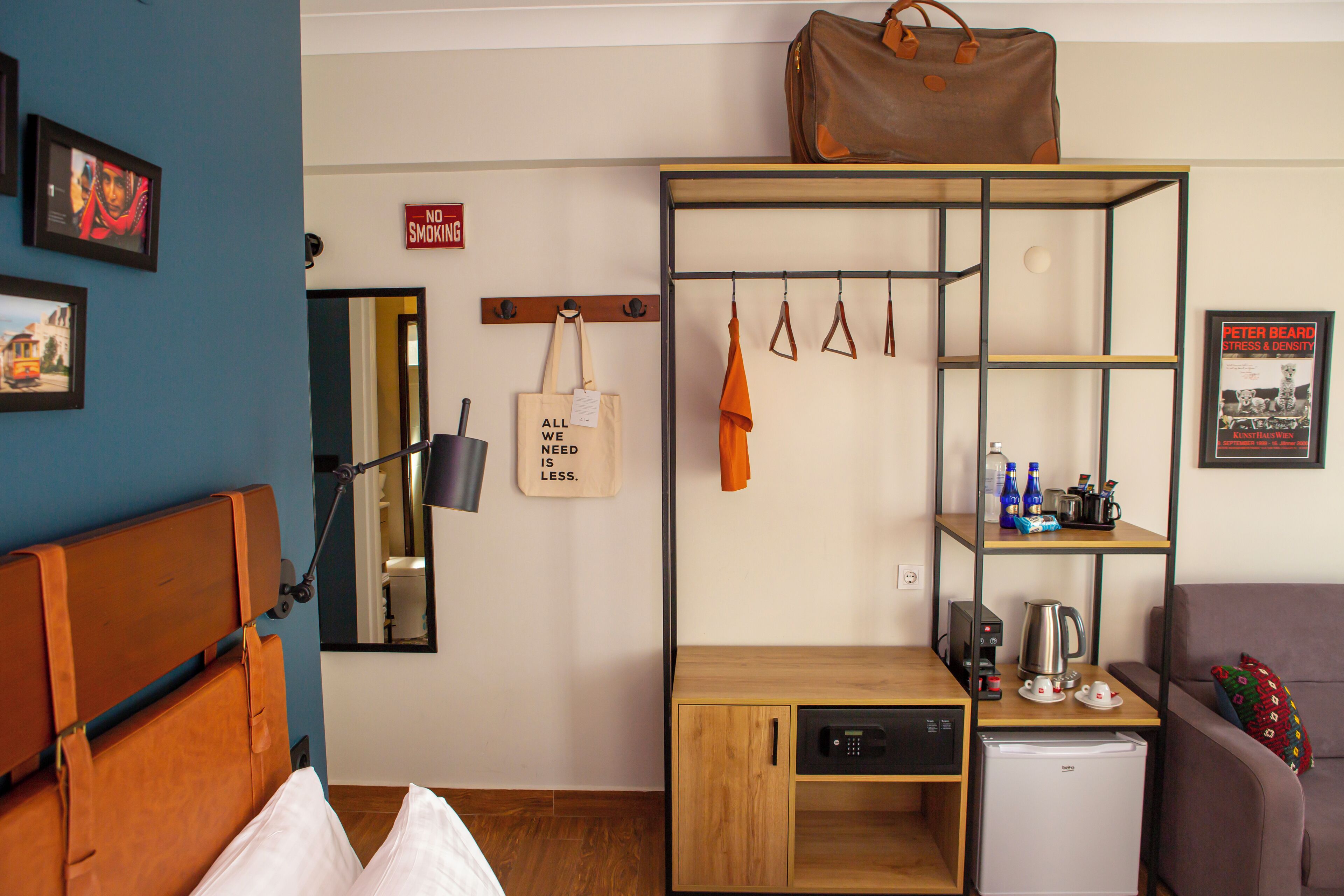 Habitación de hotel compacta con cama, escritorio con cafetera, minibar, estante con artículos de aseo, bolso colgado y cartel de 'No Fumar'.