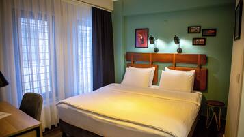 Quarto de hotel com cama, escrivaninha, cadeira e janela com cortinas.