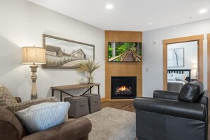 A living room with a fireplace, a television mounted on the wall, a sofa, and a coffee table. - Romantic Getaway with Private Patio and Hot Tub on Main Street - Maple Suite (Berlin)