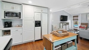 A compact kitchen with white cabinets, a microwave, and a coffee maker. There's a wooden table with a basket and a light blue cabinet below it. - Tutti Frutti (Santa Rosa Beach)