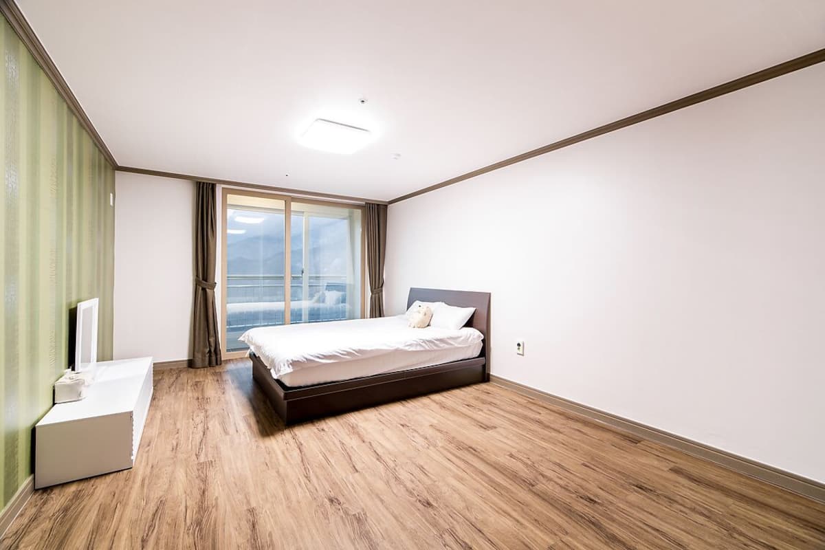A modern bedroom with a wooden floor, a bed with white bedding, a bedside table, and a large window with curtains.