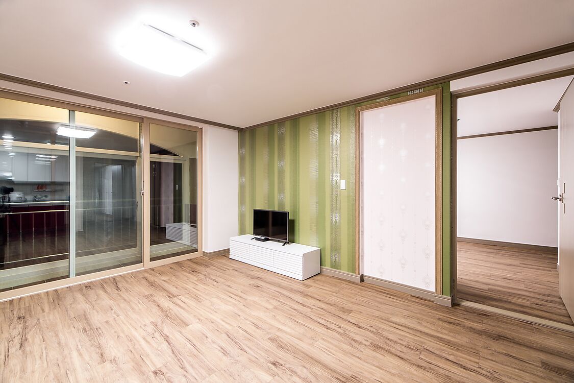A modern room with a wooden floor, a white entertainment unit, a green-striped wall, and a sliding glass door leading to a kitchen area.