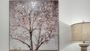 A wooden console table with a vase of dried flowers, a lamp with a patterned base, and a framed picture of a tree on the wall.