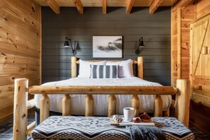 A wooden cabin bedroom with a bed, two wall-mounted lamps, a framed picture, and a small table with a cup and pastries. - Updated Cabin | Amazing Views,Hot Tub,Location (Sevierville)