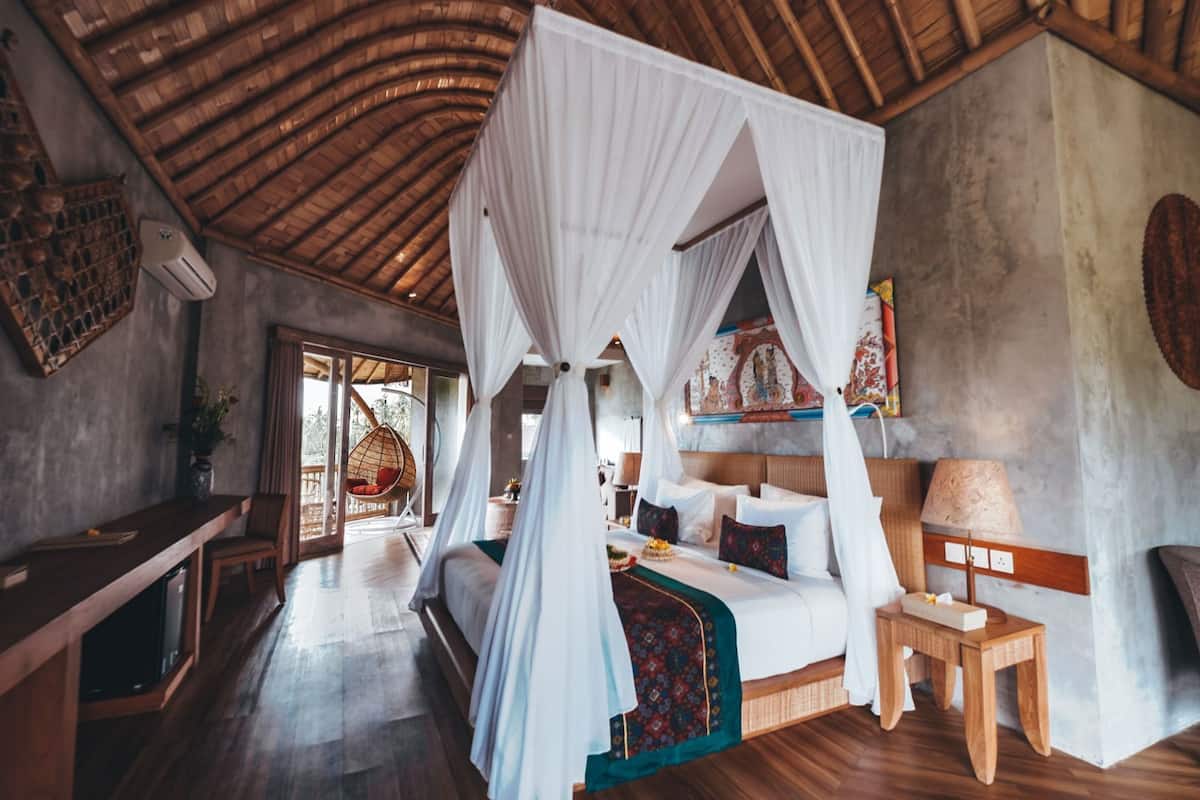 A room with a bed under a canopy, a hanging chair, a small table, and a view of the outdoors.