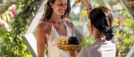 A person in a white dress holding a plate with food and a drink, standing under a structure with greenery.
