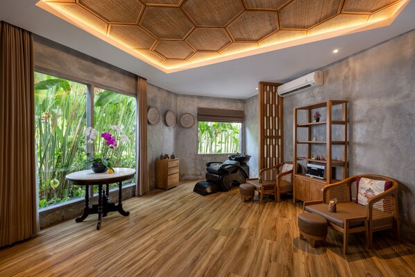 Una habitación con piso de madera, una mesa redonda con una planta, una silla de masajes, un sofá y una estantería.