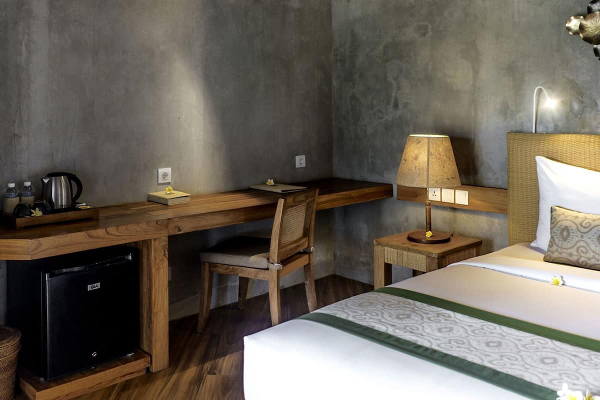 A compact hotel room with a wooden desk, a single bed, a small bedside table with a lamp, a chair, and a compact refrigerator.