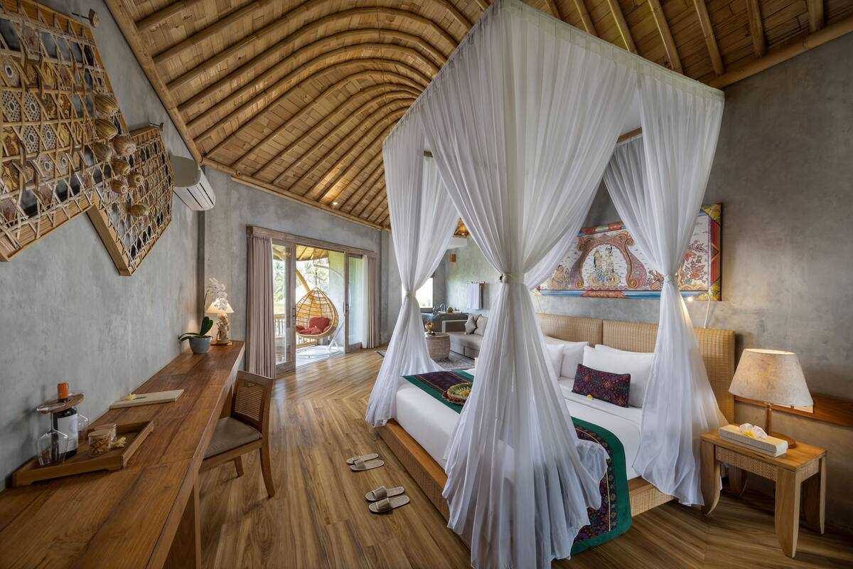 A room with a wooden floor, a bed with a canopy, a desk with a chair, and a small table with a lamp.