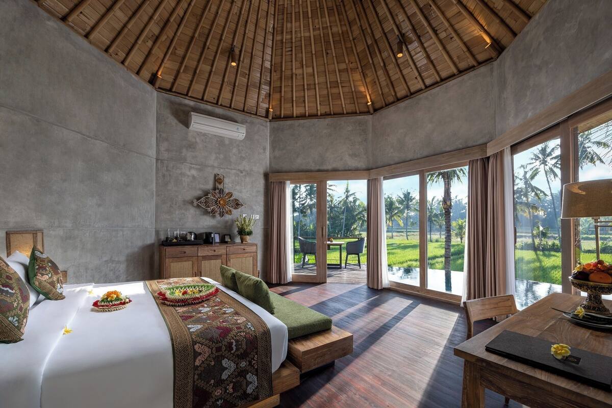 A spacious bedroom with a large bed, a wooden ceiling, and a view of a lush garden through large windows.
