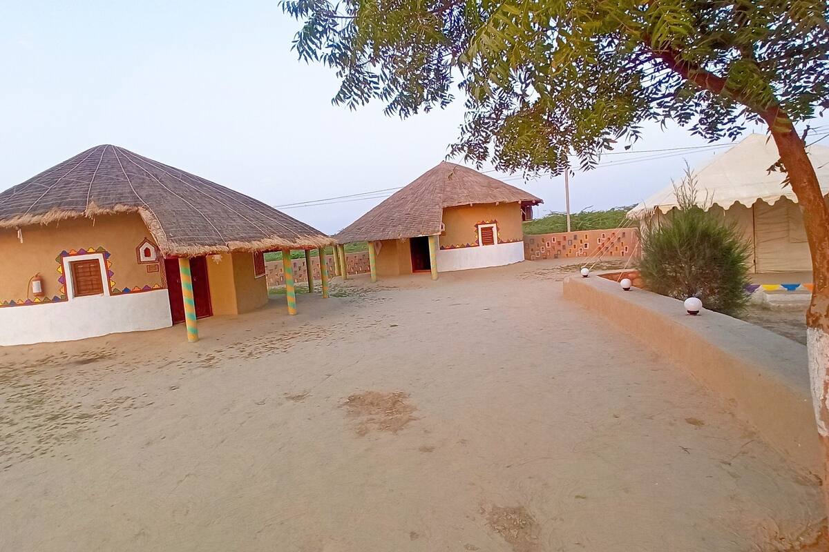 A sandy area with two thatched-roof huts, each with a door and window, and a small tree nearby.