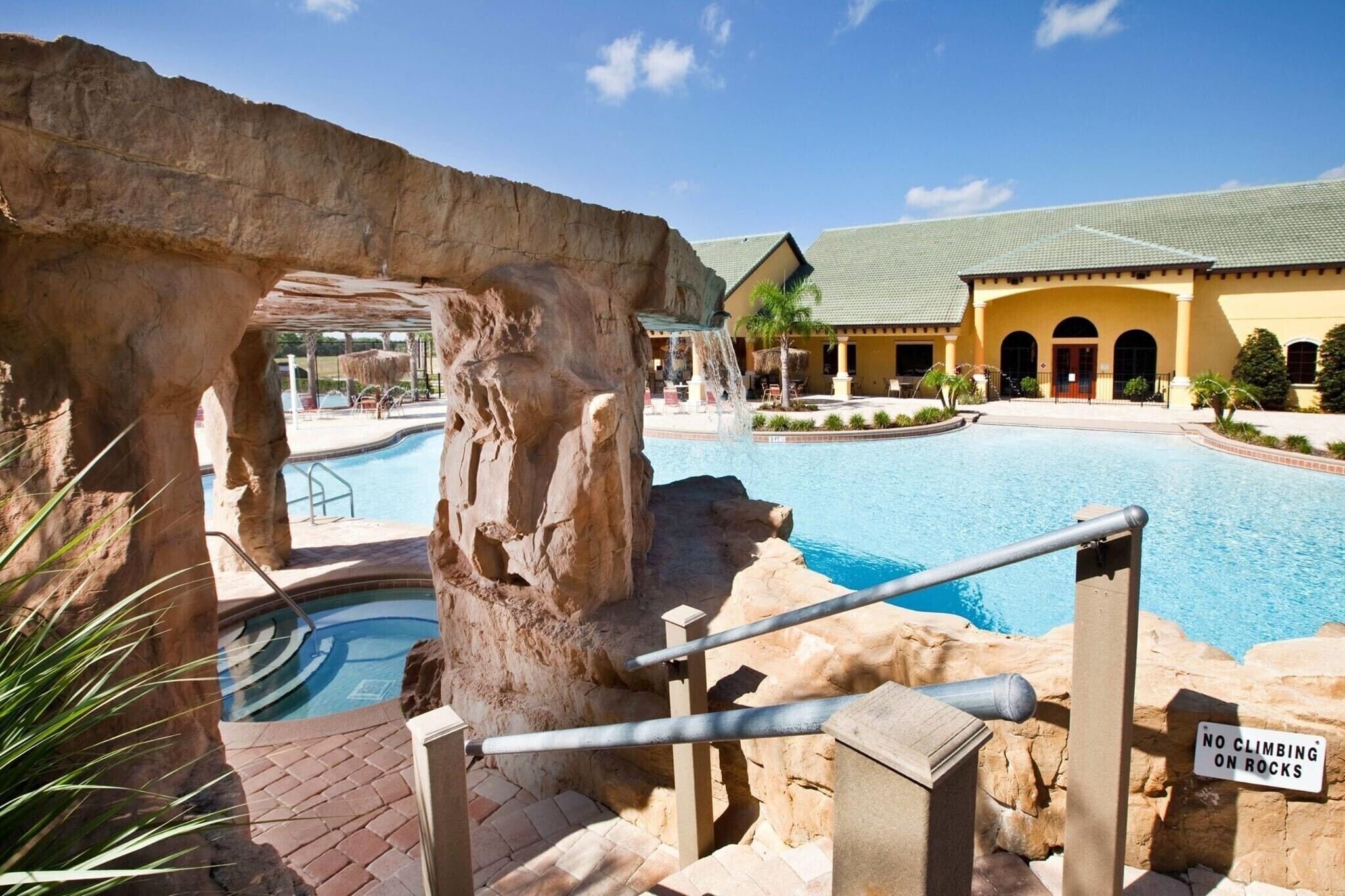 A pool area with a rock formation, a 'No Climbing on Rocks' sign, and a building with a green roof in the background.