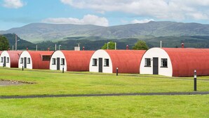 Una hilera de cabañas blancas de un piso con techos rojos, puertas y ventanas negras, frente a un paisaje de colinas verdes y un cielo despejado.