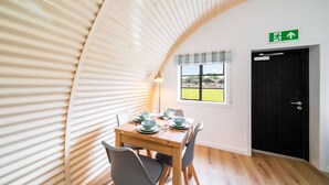 Un área de comedor con una mesa dispuesta para cuatro, una puerta negra con letrero de salida de emergencia y una ventana con vistas a un campo verde.