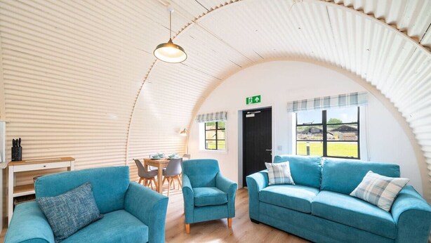 A spacious room with a curved ceiling, a dining area with a table and chairs, and a large window offering a view of the outdoors. - Hut 35 at Cultybraggan Camp (Cultybraggan Camp, Comrie)