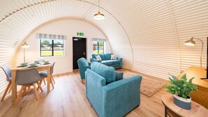 A cozy, semi-circular room with a wooden floor, a dining table set with plates and glasses, two blue sofas, and a small table with a plant. - Hut 35 at Cultybraggan Camp (Cultybraggan Camp, Comrie)