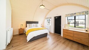 A bedroom with a curved ceiling, a bed with a white and yellow bedspread, a wooden dresser, and a window with a view of a building outside. - Hut 35 at Cultybraggan Camp (Cultybraggan Camp, Comrie)