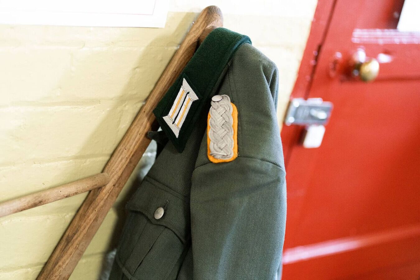 A green jacket with a patch and a metal button, hanging on a wooden hanger against a wall.
