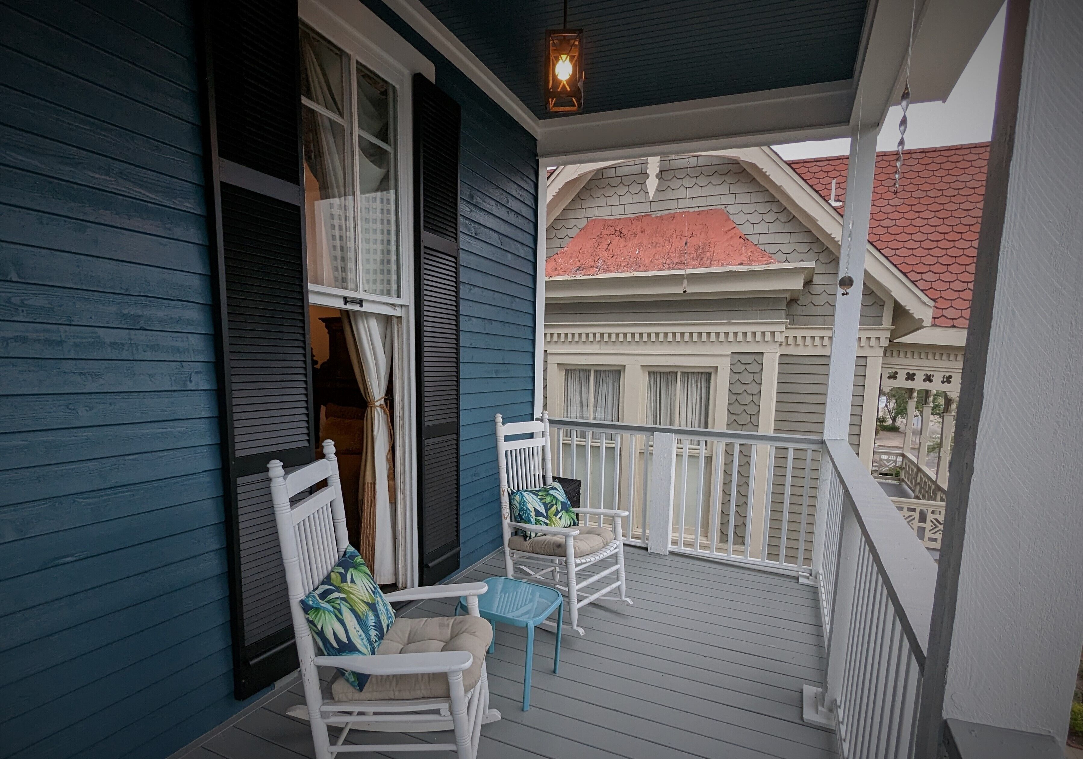 A covered porch with two white rocking chairs and a small table, overlooking a building with a red roof.