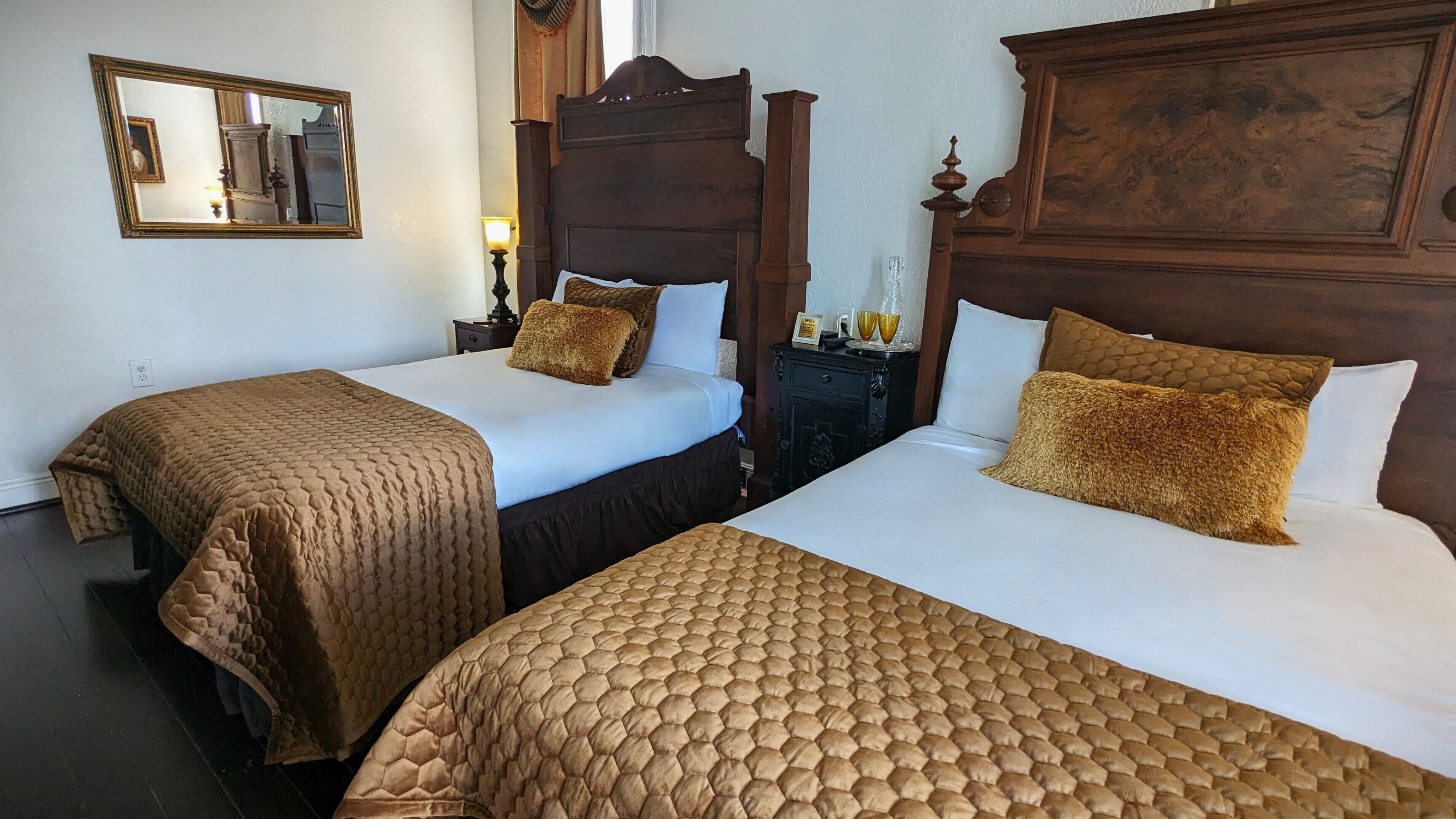 Ein Hotelzimmer mit zwei Einzelbetten, jeweils mit brauner, gesteppter Bettdecke und passenden Kissen. Jedes Bett hat ein Holz-Kopfbrett, und dazwischen befindet sich ein Nachttisch mit Lampe und einem gerahmten Bild.