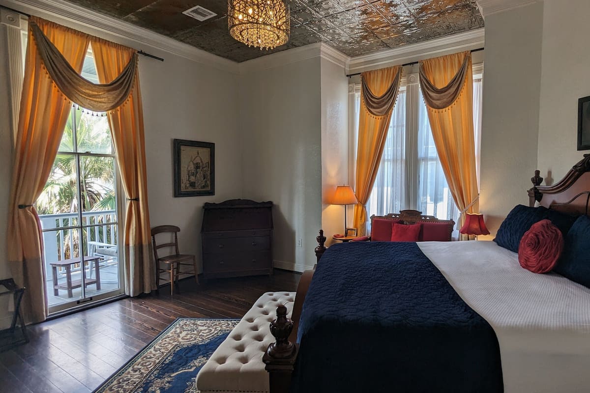 A hotel room with a large bed, a wooden dresser, a chair, a balcony, and a chandelier.