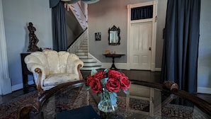 A grand room with a classic armchair, a glass coffee table with a book and flowers, a staircase, and a door.