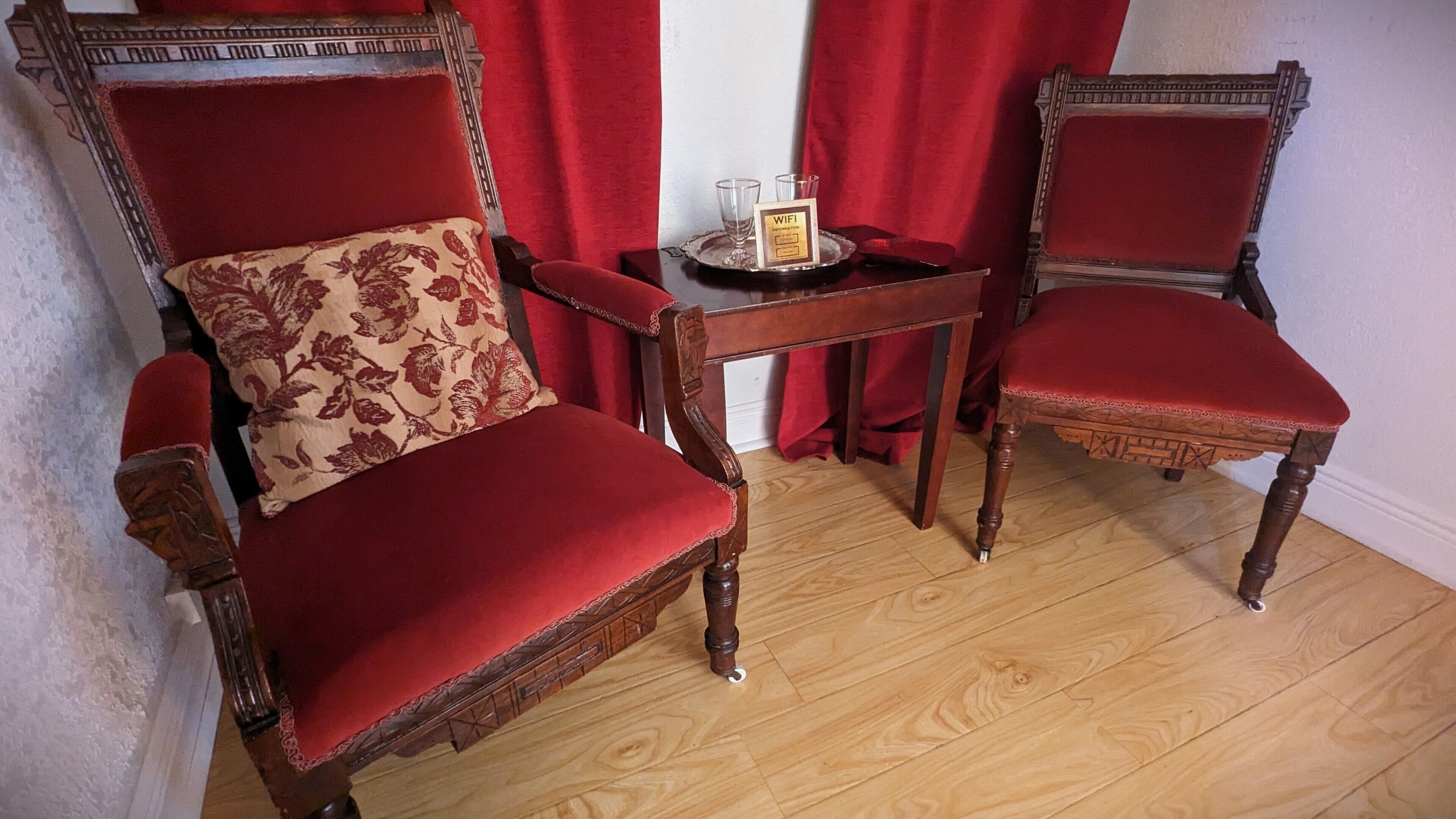Duas cadeiras de madeira ornamentada com estofamento vermelho e uma pequena mesa com um tabuleiro que contém um copo e uma placa.