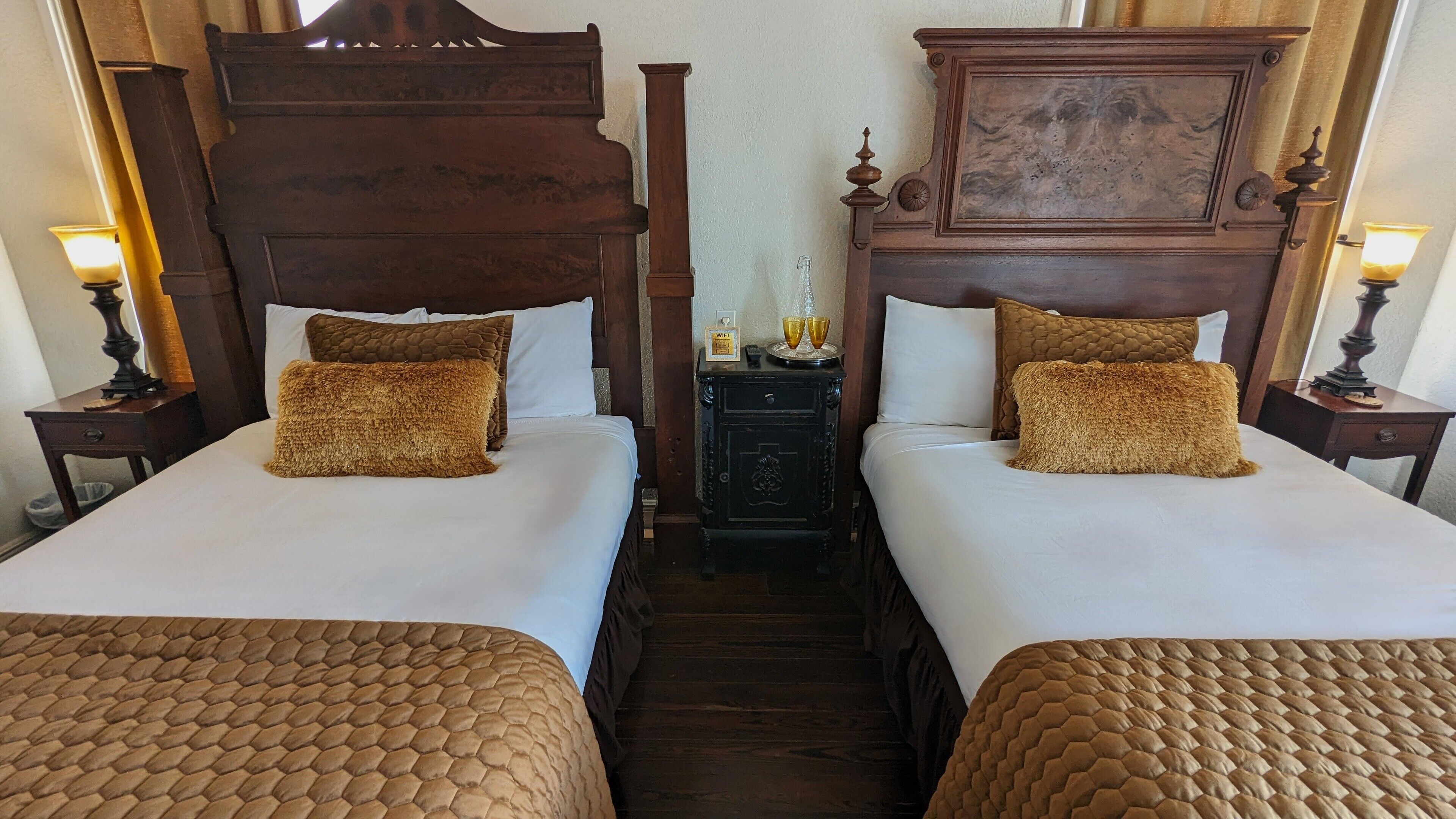 A hotel room with two beds, each with a wooden headboard and a brown quilted bedspread. There is a small wooden nightstand between the beds with a lamp and a small table with a glass and two cups.