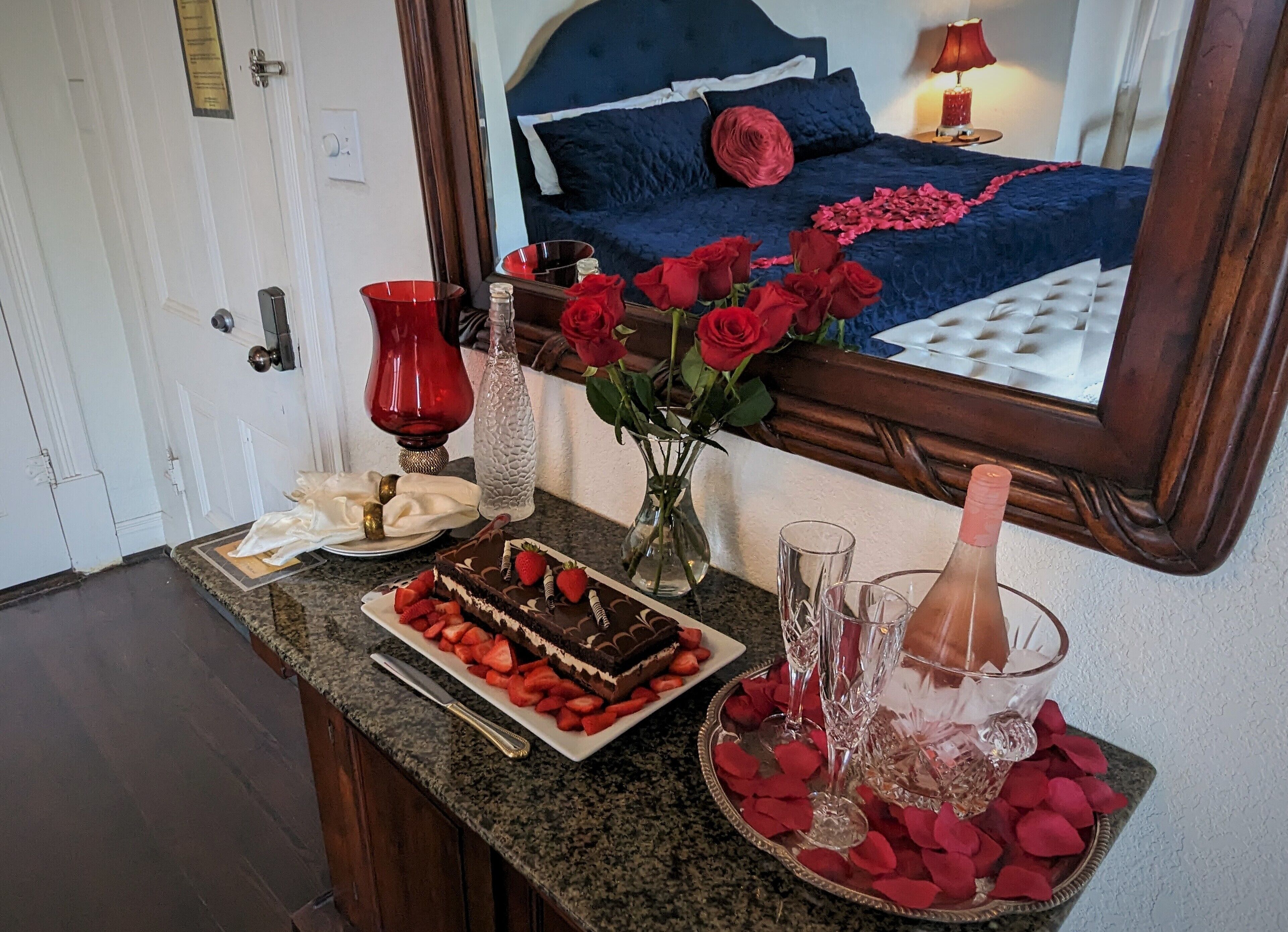 Une chambre avec un lit, un miroir, une table de chevet, un vase rempli de roses rouges, un plateau avec des fraises et un gâteau au chocolat, et une bougie rouge.