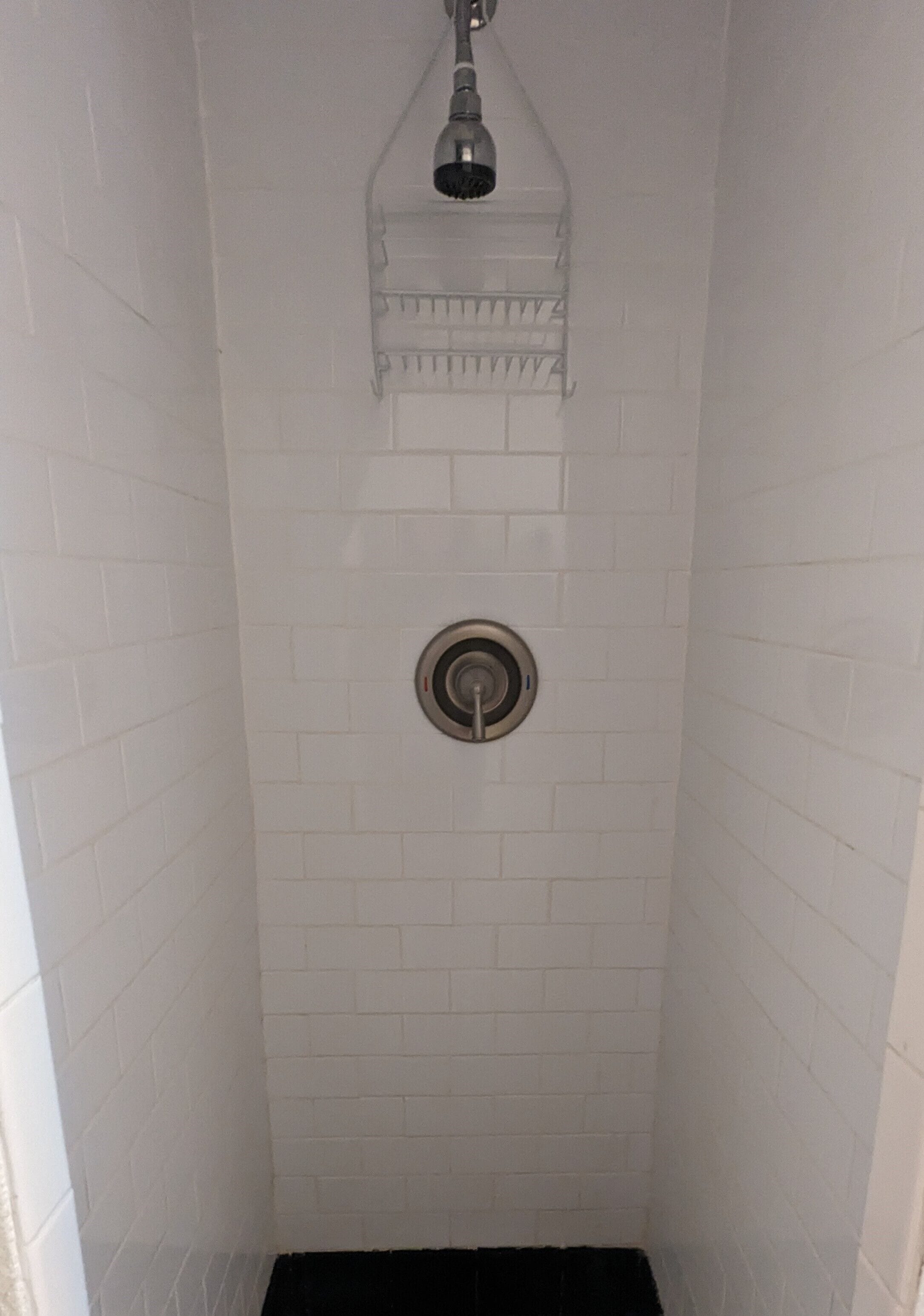 A narrow shower stall with white tiled walls and a black drain.
