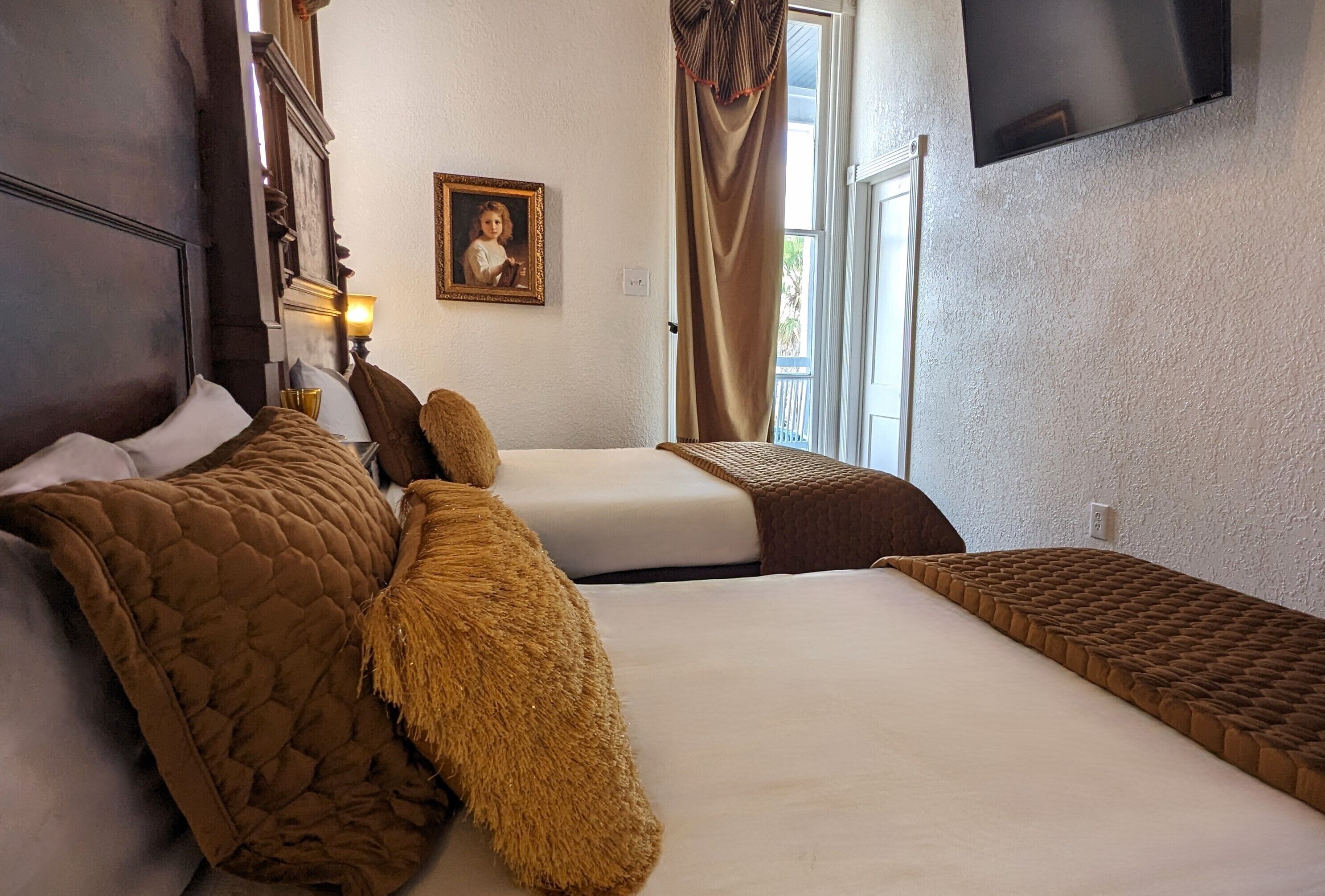 A hotel room with two beds, a television mounted on the wall, a framed picture on the wall, and a window with curtains.