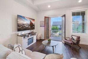 Una sala de estar con un televisor de pantalla plana grande, un sofá, una mesa de centro y una silla. Hay una puerta de vidrio corredera que da a un balcón con vistas al barrio.