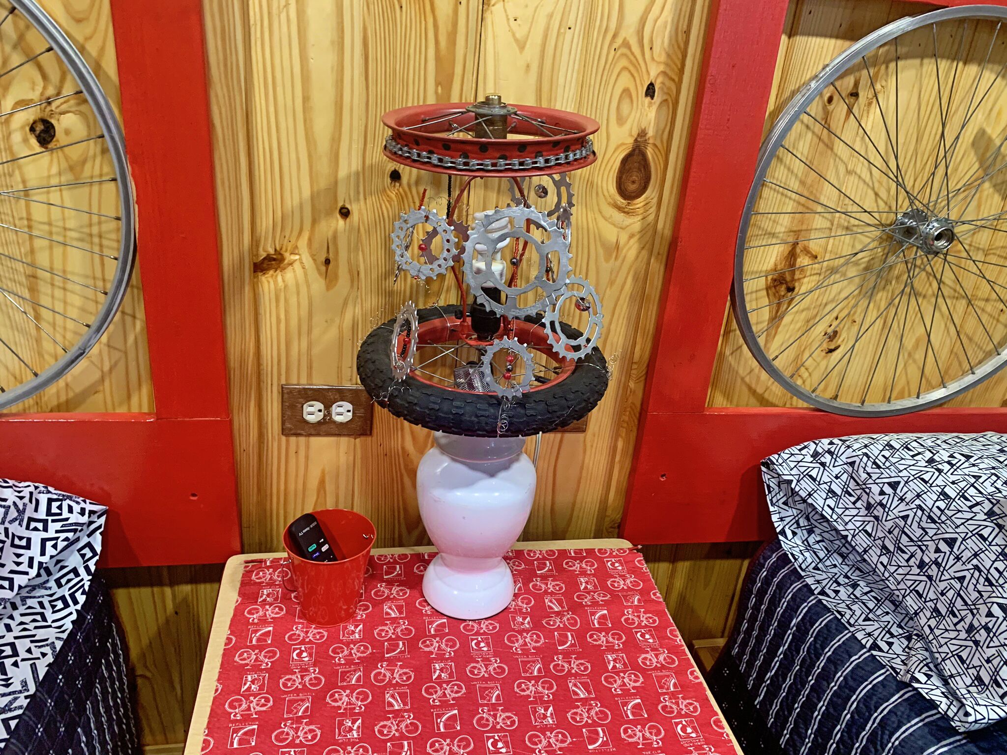 Een kamer met een houten wand, een fietswiel, een wit voetstuk met tandwielen, een rode tafel met een fietsmotief, en een rode emmer met een afstandsbediening.