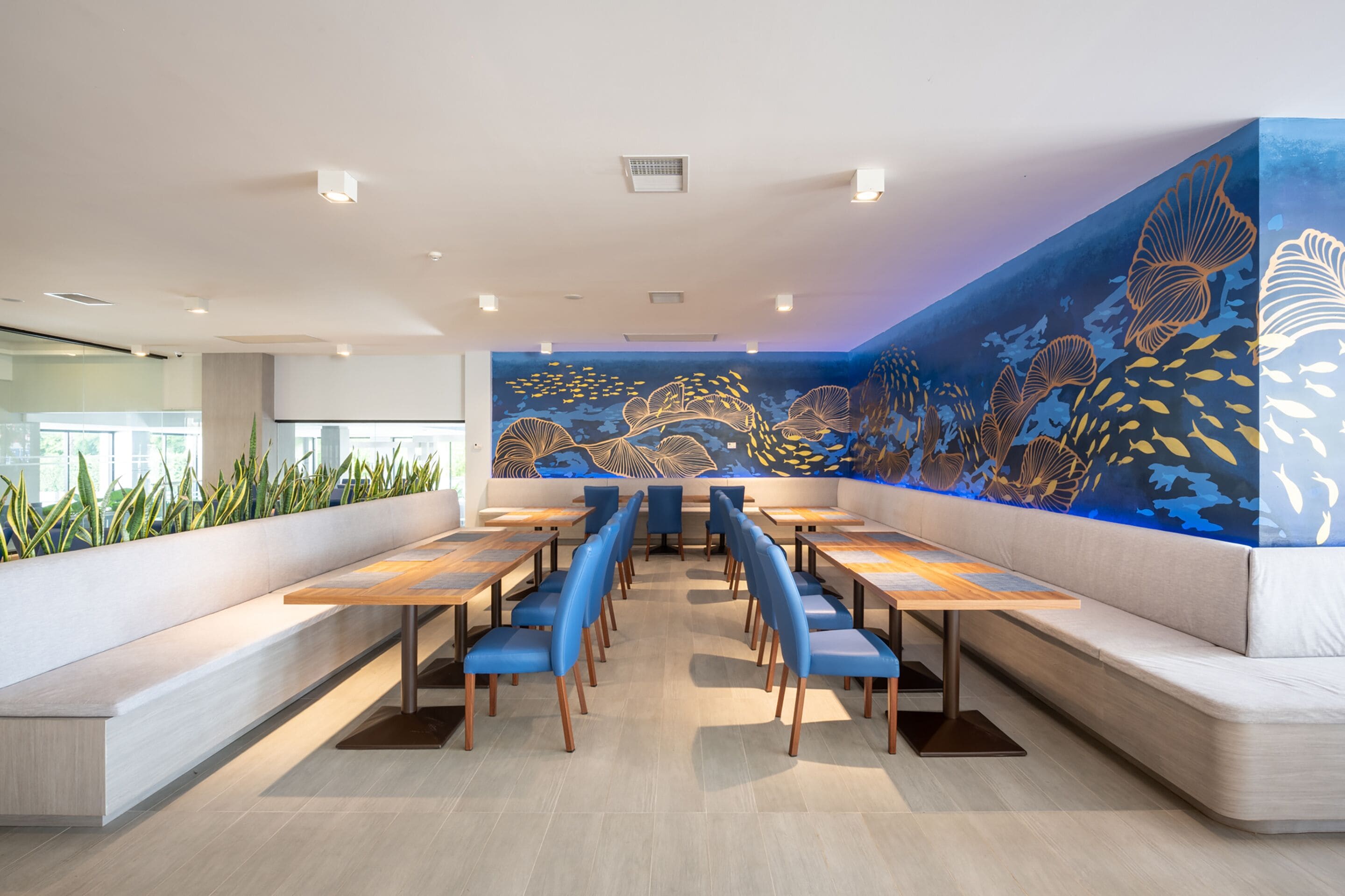 A modern restaurant interior with long wooden tables, blue cushioned chairs, and a large mural of marine life on the back wall.